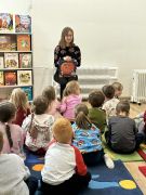 Dzieci w bibliotece siedzą na dywanie słuchają opowiadania. Pani czyta.